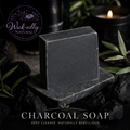 A rectangular bar of charcoal soap displayed on a dark stone slab, surrounded by black charcoal pieces, green leaves, and a lit candle in the background. The 'Wick-edly Natural' brand logo is visible in the top-left corner, and the text 'CHARCOAL SOAP' is displayed at the bottom.